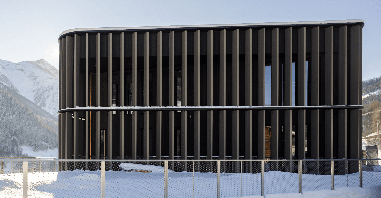 Modern two-storey building with dark vertical timber fins and rounded corners, snow on ledges, set against alpine mountains.