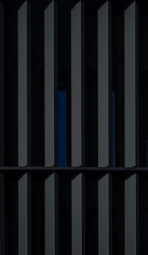 Close-up view of a modern building façade with vertical dark metal fins, creating a repeating geometric pattern with narrow gaps that reveal a deep blue background.
